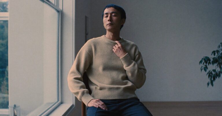 A person sitting upright beside a window with one hand resting on the chest, mid-exhale during a physiological sigh